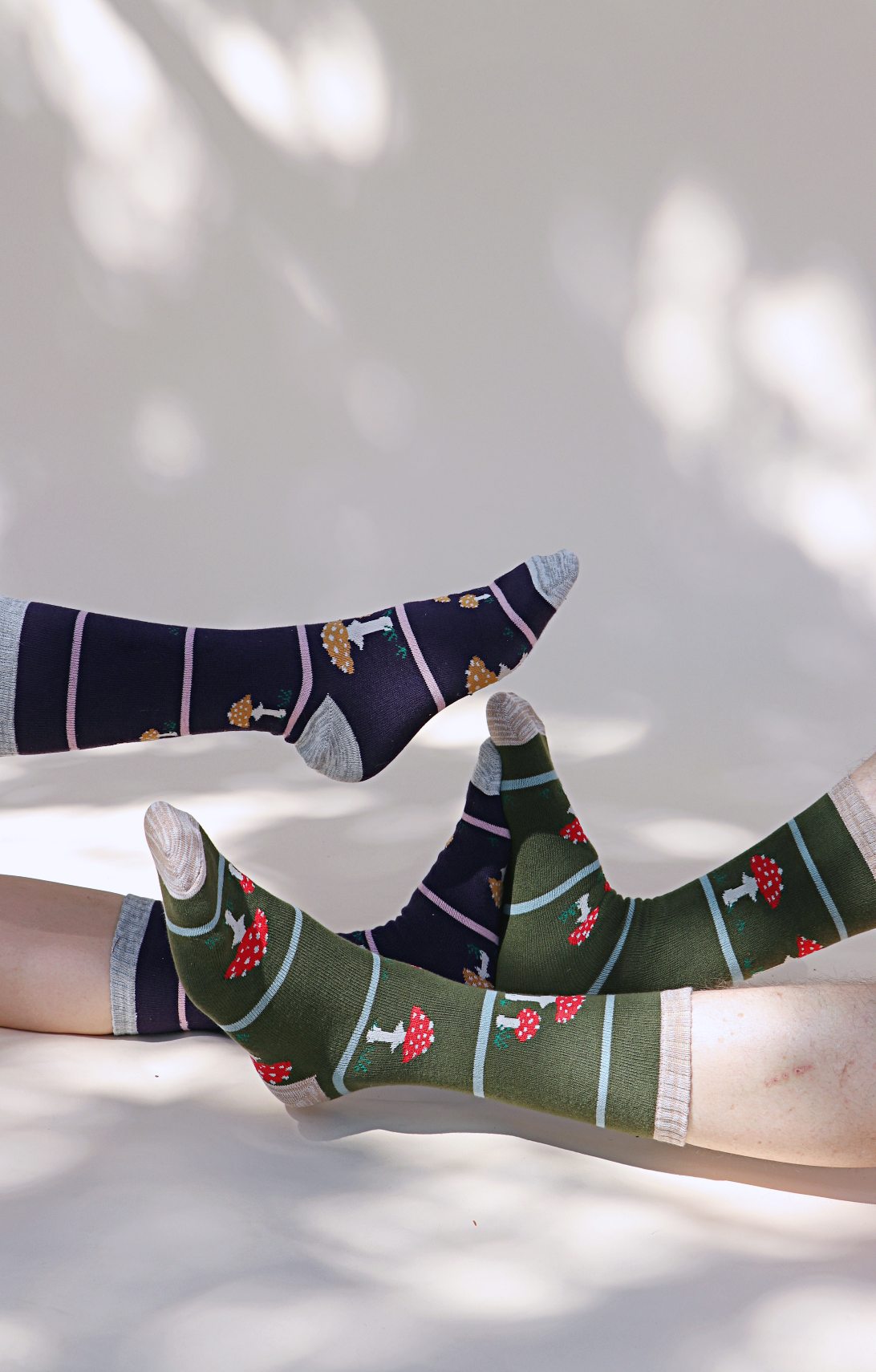 TABBISOCKS brand Replant Pairs Mushroom Socks in green Olive color with red mushroom design with white dots all over and ivory point color at the toes and cuffs of the socks; legs of a man wearing Grey half pants and a woman wearing socks in purple with brown mushroom design with Grey point color all over.