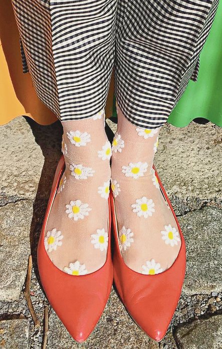 Leg of a woman wearing white and black gingham check pants with orange pumps wearing socks in CLEAR WHITE color of a product called Daisy Sheer Socks of the TABBISOCKS brand with white daisy flowers illustrated throughout on a transparent white fabric