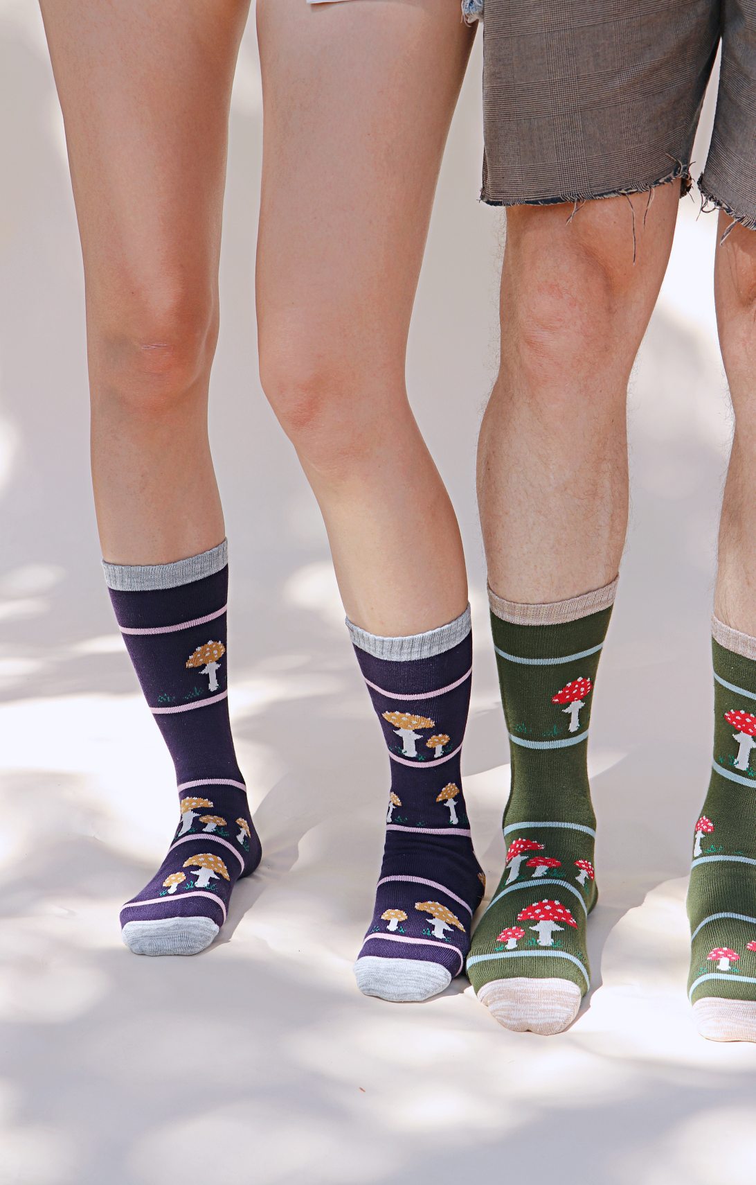 TABBISOCKS brand Replant Pairs Mushroom Socks in green Olive color with red mushroom design with white dots all over and ivory point color at the toe and mouth of the socks, lower body of a man wearing Grey half pants and a woman wearing socks in purple with brown mushroom design with Grey point color all over.