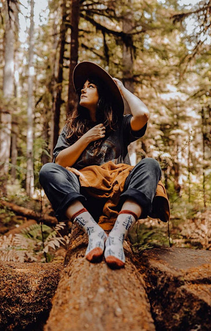 A woman sitting in a forest wearing TABBISOCKS brand Replant Pairs Tree Socks in Beige with brown inserts at the cuff and toe and an overall tree design