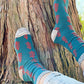 A woman is in the forest wearing TABBISOCKS brand Replant Pairs Tree Socks in Teal with Grey at the cuff and toe and a reddish brown tree design throughout the socks