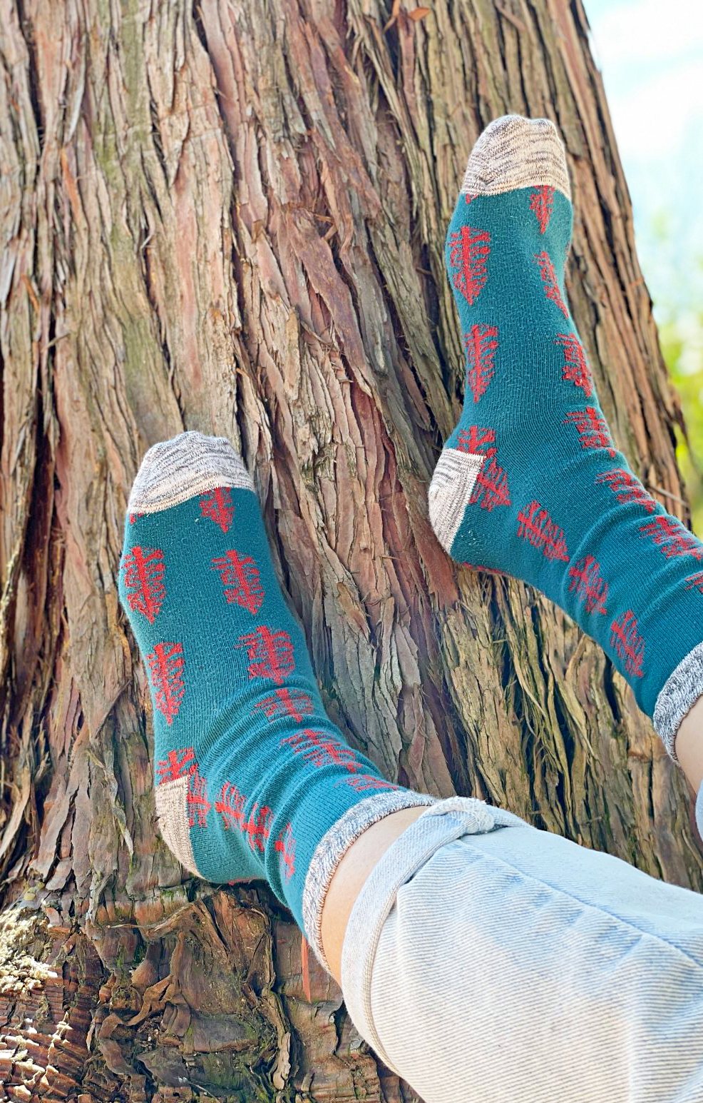 A woman is in the forest wearing TABBISOCKS brand Replant Pairs Tree Socks in Teal with Grey at the cuff and toe and a reddish brown tree design throughout the socks