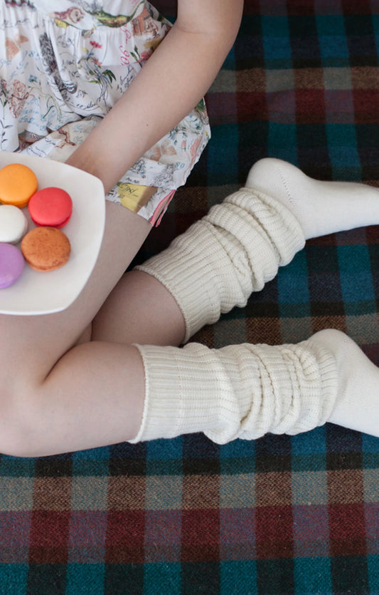 A woman wearing TABBISOCKS brand Scrunchy Over the Knee Socks, knee-length knee socks in Ivory color, relaxing while eating macarons.