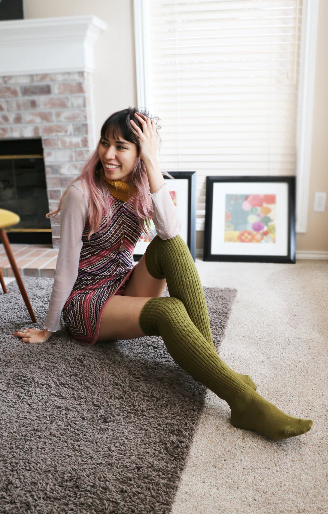 A pink-haired woman relaxing in her living room wearing TABBISOCKS brand Scrunchy Over the Knee Socks, knee-length knee socks in Olive color