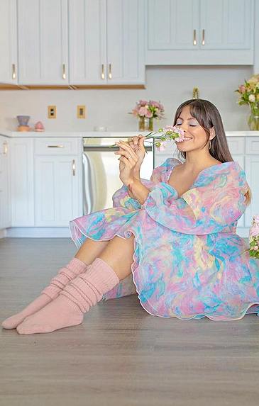 A woman wearing a colorful lace dress sitting in the dining room smelling flowers wearing TABBISOCKS brand Scrunchy Over the Knee Socks, a pale pinkish Petal color of knee-length knee socks