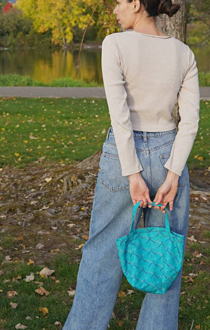 Person holding a turquoise woven handbag in an outdoor setting with greenery.