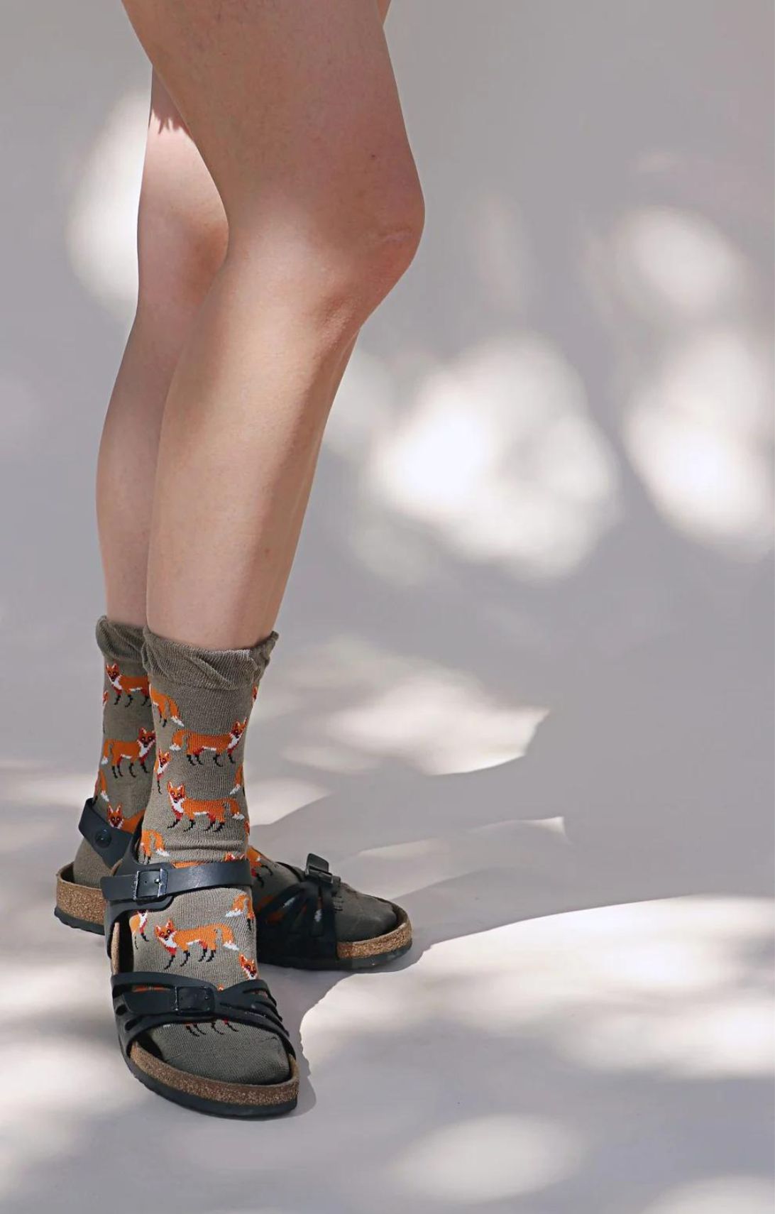 A person's foot wearing a pair of TABBISOCKS brand Animal Rescue Pairs Fox Socks, which are OLIVE BEIGE in color with an orange fox illustration throughout, with a pair of black sandals and a standing person's foot