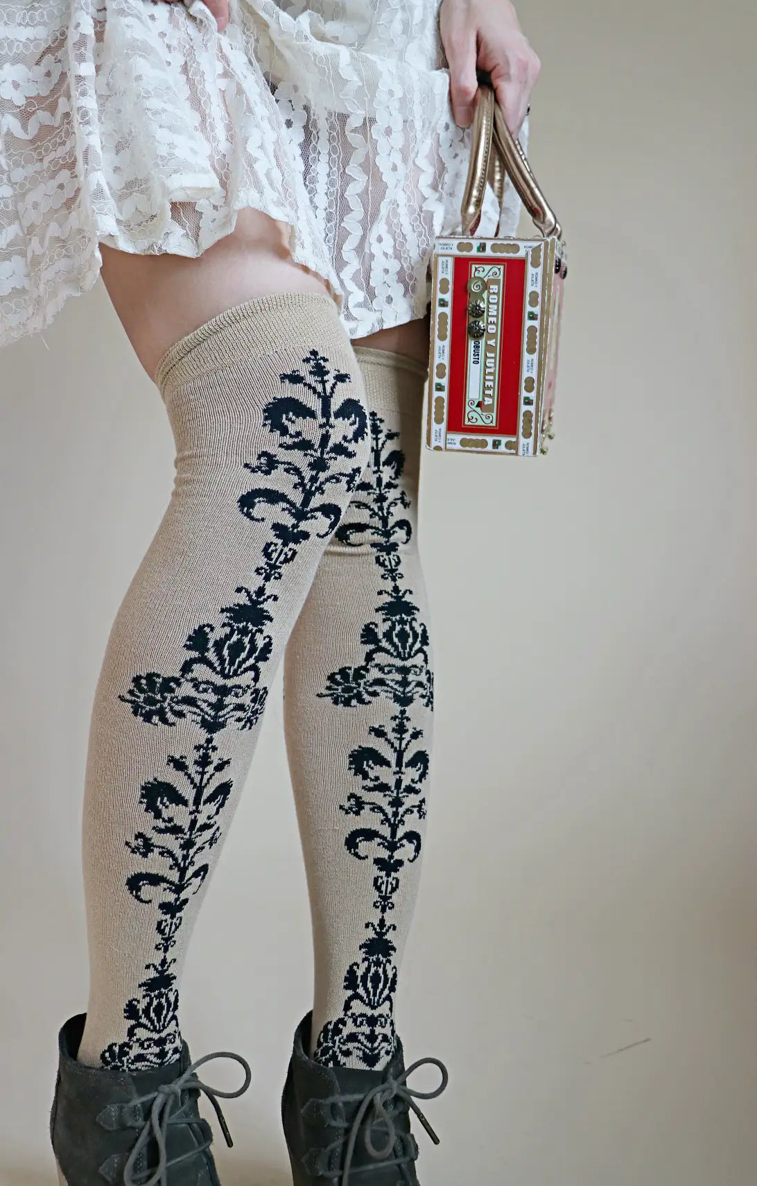 Lower half of a woman wearing TABBISOCKS brand Floral Chain Over The Knee Socks, knee-length socks in beige with a black floral pattern on the front center portion, worn with a white lace skirt and suede shoes