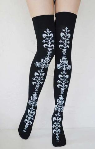 Female leg wearing TABBISOCKS brand Floral Chain Over The Knee Socks, a pair of knee-length socks in black with a white floral pattern on the front center portion