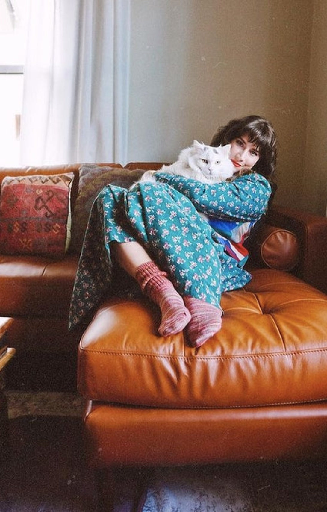A woman sitting on a leather couch with a Persian cat in a pastel green dress wearing Rose Mix color of TABBISOCKS brand Lounge Scrunchy Crew Socks