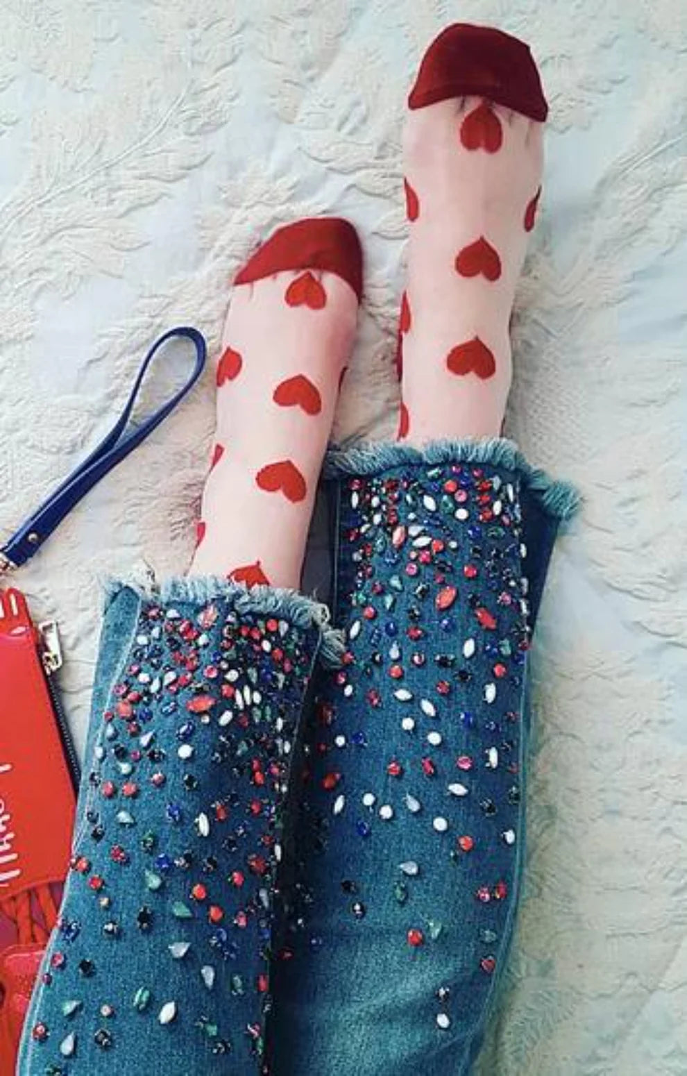 Leg of a woman wearing jeans with a red color of a product called Black Heart Sheer Socks of the TABBISOCKS brand, a transparent transparent fabric with a red heart pattern all over it