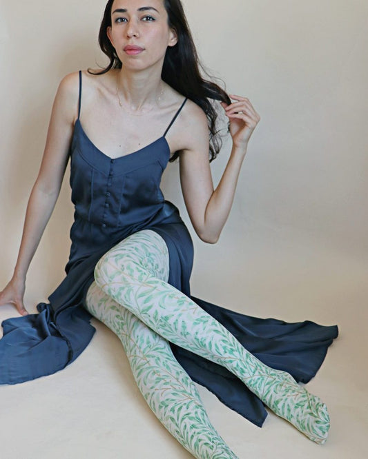 Tights called Willow Boughs Green from the William Morris collection of the TABBISOCKS brand, with an overall design of green plants, the color antique-looking green, worn by a woman in a navy silk dress, sitting